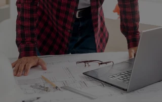 Oberkörper einer Person in kariertem Hemd über einem Tisch mit technischen Zeichnungen, Brille und aufgeklapptem Laptop