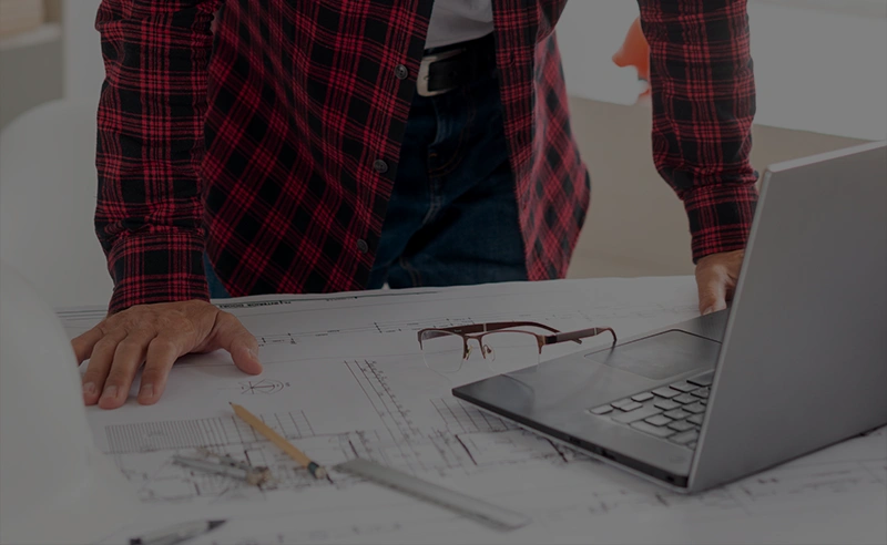 Oberkörper einer Person in kariertem Hemd über einem Tisch mit technischen Zeichnungen, Brille und aufgeklapptem Laptop